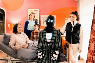 Eine Gruppe von Personen steht in einem Schlafzimmer mit einer älteren Frau im Bett. Ein Roboter in einem Bademantel steht neben ihnen und fügt sich in die Szene ein.
