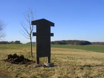 Eine schwarze Informationstafel steht auf einer Wiese vor einem klaren Himmel. Im Hintergrund sind Bäume und ein grünes Feld zu sehen.