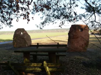 Zwei große Steine stehen auf einer Wiese, zwischen ihnen ist eine Holzbank mit Tisch. Im Hintergrund erstreckt sich eine grüne Landschaft unter einem klaren Himmel.