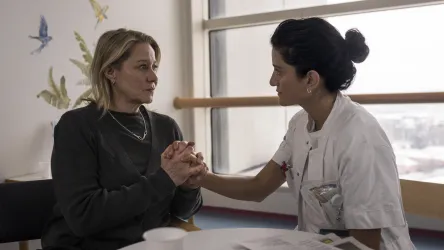 Eine Frau und eine Pflegekraft sitzen sich gegenüber und halten sich an den Händen. Die Atmosphäre ist einfühlsam und unterstützend in einem medizinischen Umfeld.