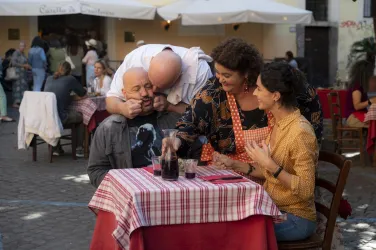 Eine lebhafte Restaurantszene im Freien mit mehreren Personen. Zwei Frauen interagieren fröhlich mit zwei Männern am Tisch.