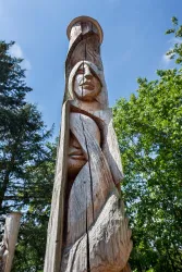 Ein kunstvoll geschnitzter Totempfahl aus Holz zeigt menschliche Gesichter. Der Totempfahl steht im Freien, umgeben von Bäumen und blauem Himmel.