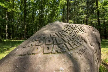 Ein großer Stein im Wald mit eingravierten Worten. Die Umgebung ist grün und natürlich, umgeben von Bäumen.