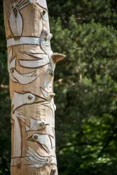Ein kunstvoll geschnitzter Baumstamm mit Vogeldarstellungen. Im Hintergrund sind grüne Bäume sichtbar.