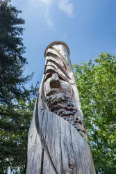 Ein kunstvoll geschnitzter Holztotem, der die Form eines Gesichts zeigt. Der Totem steht im Freien, umgeben von Bäumen und blauem Himmel.