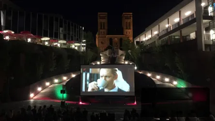 Ein Freiluft-Kino zeigt einen Film auf einer großen Leinwand. Die Kulisse besteht aus stilvollen Gebäuden und einer stimmungsvollen nächtlichen Atmosphäre.