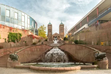 Ein schöner Brunnen mit Wasserfällen und Schwanenfiguren im Vordergrund. Im Hintergrund sind zwei markante Türme einer Kirche sichtbar.