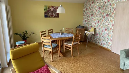 Ein helles Esszimmer mit einem runden Tisch und Stühlen. Die Wände sind mit blühendem Muster tapeziert und ein gemütlicher Sessel steht in der Ecke.