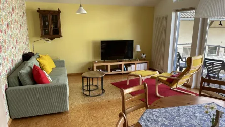 Ein helles Wohnzimmer mit einem grünen Sofa und einem Fernseher. Es gibt einen Tisch, Stühle und großen Fenstern, die viel Tageslicht hereinlassen.