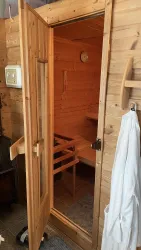 Eine hölzerne Sauna mit offenem Eingang. Innen sind eine Holzbank und ein Thermostat sichtbar.