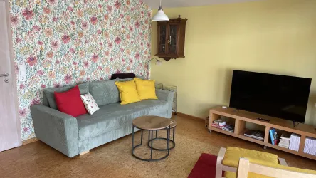 Ein modernes Wohnzimmer mit einer grünen Couch, bunten Kissen und einem runden Tisch. An der Wand ist eine Blumenmuster-Tapete und ein Fernseher auf einem Regal.