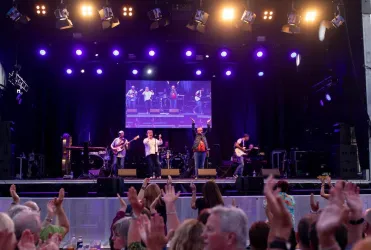 Ein Bild eines bunt beleuchteten Konzertes, mit Bühne auf der eine Band spielt. Im Vordergrund sieht man eine Gruppe von Menschen die zur Musik tanzen.