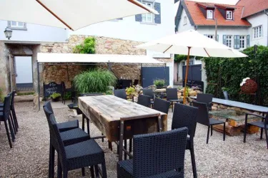 Ein gemütlicher Außenbereich mit einem großen Tisch und schwarzen Stühlen. Sonnenschirme bieten Schatten, umgeben von grünen Pflanzen und einer rustikalen Steinmauer.