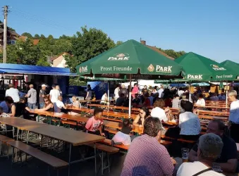 Ein belebtes Festival mit vielen Menschen, die an langen Holztischen sitzen. Grüne Sonnenschirme bieten Schatten an einem sonnigen Tag.