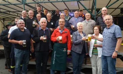 Eine Gruppe von Menschen steht zusammen und hebt Bierkrüge. Sie lächeln und scheinen eine festliche Stimmung zu genießen.