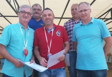 Eine Gruppe von fünf Männern steht zusammen und lächelt in einer hellen Umgebung. Sie halten Dokumente in der Hand und wirken zufrieden.