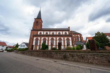 Eine alte Kirche mit einem hohen Turm und bunten Fenstern. Im Hintergrund sind weitere Gebäude und eine bewölkte Himmel zu sehen.