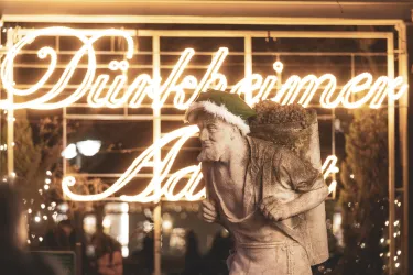 Eine Statue eines Mannes mit einem Weihnachtsmann-Hut und einem Sack auf dem Rücken. Im Hintergrund leuchtet ein festliches Schild mit dem Schriftzug „Dürkheimer Advent“.