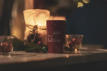 Eine Tasse mit dem Aufdruck „Advent“ steht auf einem Tisch. Im Hintergrund sind ein sanft leuchtendes Licht und dekorative Gläser zu sehen.