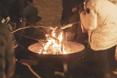 Ein Lagerfeuer, umgeben von Menschen, die Stockbrot rösten. Die Flammen leuchten warm in der Dunkelheit.