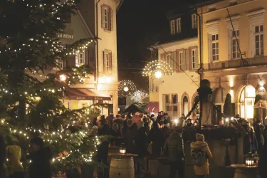 Ein festlicher Weihnachtsmarkt bei Nacht mit vielen Lichtern und fröhlichen Menschen. Im Vordergrund steht ein Weihnachtsbaum, umgeben von bunten Ständen.