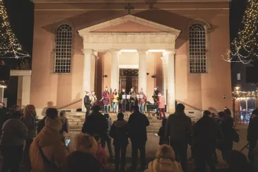 Eine Gruppe von Sängern steht auf einer Treppe vor einer Kirche, während viele Zuschauer zuhören. Die Szene ist festlich mit Lichtern dekoriert und es ist Abend.