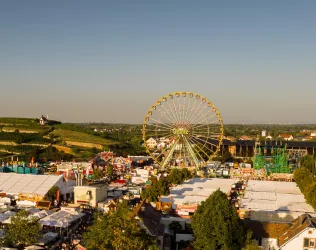Ein großer Jahrmarkt mit einem Riesenrad und vielen bunten Ständen. Die Umgebung ist grün und die Atmosphäre festlich.