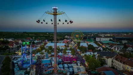 Eine farbenfrohe Kirmes mit verschiedenen Fahrgeschäften und Lichtern. Im Hintergrund sind ein Riesenrad und ein großer Türmchen sichtbar.