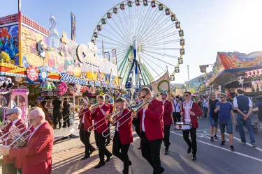 Eine lebhafte Straßenparade mit Musikern in roten Jacken, die Trompeten und Posaunen spielen. Im Hintergrund ist ein großes Riesenrad und festliche Attraktionen zu sehen.