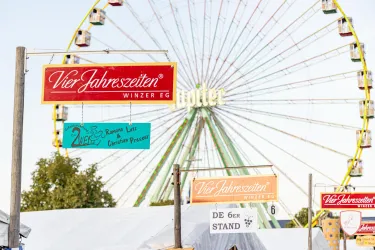 Ein Jahrmarkt mit einem großen Riesenrad im Hintergrund. Verschiedene Schilder präsentieren Stände und Attraktionen.