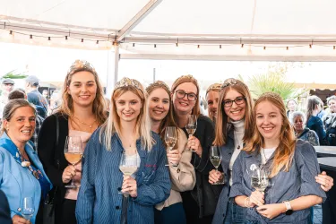 Eine Gruppe junger Frauen posiert lächelnd unter einem Zelt, jede mit einem Glas in der Hand. Sie tragen Kleider und Krönchen, die eine festliche Atmosphäre schaffen.