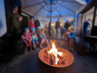 Ein warmes Lagerfeuer in einer gemütlichen Umgebung. Im Hintergrund sitzen Menschen, die die Atmosphäre genießen.
