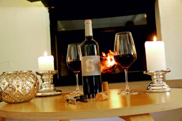 Ein Weinflasche steht auf einem Tisch neben zwei Weingläsern und Kerzen. Im Hintergrund flackert ein Kaminfeuer.
