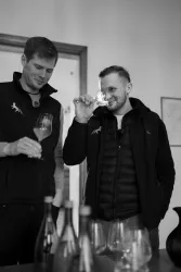 Zwei Männer halten Gläser in der Hand und riechen daran. Im Hintergrund stehen Weine auf einem Tisch.