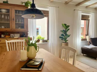 Ein gemütlicher Essbereich mit einem Holztisch, auf dem eine Blumenvase mit Tulpen steht. Große Fenster lassen viel Tageslicht hereinfällt und sorgen für eine helle Atmosphäre.