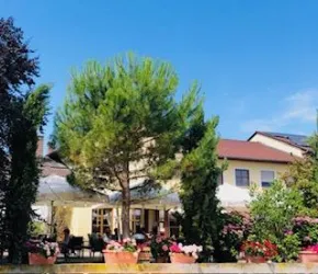 Ein gemütliches Restaurant mit einem großen Baum und vielen Blumen. Unter den Sonnenschirmen sitzen Gäste und genießen die Atmosphäre.