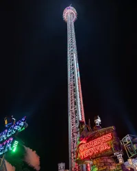 Ein hoher Fahrstuhl mit bunten Lichtern steht in einem Vergnügungspark bei Nacht. Im Vordergrund befindet sich der Schriftzug "Fortress Tower".