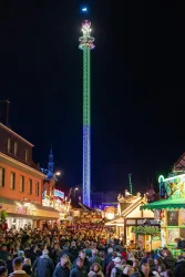 Eine belebte Weihnachtsmarktstraße bei Nacht, beleuchtet von festlichen Lichtern. Im Hintergrund ragt ein hoher, bunt beleuchteter Turm in den Himmel.
