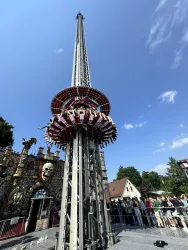 Ein hoher Freifallturm mit Fahrgästen, die nach oben geschossen werden. Im Hintergrund sind Menschen und eine dekorierte Festhalle zu sehen.