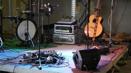 Ein Musik-Setup auf einer Bühne mit verschiedenen Instrumenten wie einer Gitarre, Schlagzeug und Mischpult. In der Umgebung gibt es viele Kabel und elektrische Geräte.