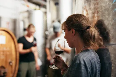 Eine Frau mit einem Glas Rotwein steht in einem Weinkeller. Im Hintergrund sind andere Personen und Weinfässer zu sehen.