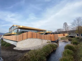 Ein modernes Gebäude mit großen Fenstern und einem goldenen Dach. In der Nähe fließt ein kleiner Bach und es sind einige Bäume und Pflanzen zu sehen.
