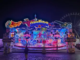 Ein bunter Jahrmarkt mit leuchtenden Lichtern und aufregenden Attraktionen. Die Szene zeigt ein Fahrgeschäft mit Themen aus berühmten Comic-Helden.