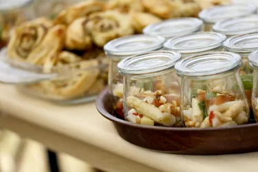 Ein Tisch mit kleinen Gläsern, die bunte Salate enthalten. Im Hintergrund sind köstliche, frisch gebackene Teigrollen zu sehen.