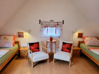 Ein gemütliches Schlafzimmer mit zwei Einzelbetten und zwei Sesseln. Das Fenster ist mit Vorhängen dekoriert und ein kleiner Tisch steht in der Mitte.