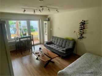 Ein modernes Wohnzimmer mit einem grauen Sofa und einem Holztisch. An der Wand hängen Weinfalschen und große Fenster bieten viel Tageslicht.