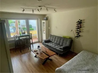 Ein modernes Wohnzimmer mit einem grauen Sofa und einem Holztisch. Große Fenster bringen viel Licht in den Raum und ein Esstisch ist ebenfalls vorhanden.