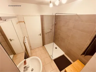 Ein modernes Badezimmer mit einer Dusche und einem Waschbecken. Die Wände sind hell gefliest und es gibt einen Holzregalboden.