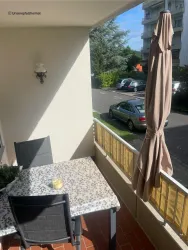 Ein gemütlicher Balkon mit einem Tisch und Stühlen. Im Hintergrund sind Autos und viel Grün sichtbar.