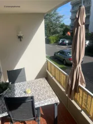 Ein schöner Balkon mit einem Tisch und Stühlen. Im Hintergrund sind Bäume und parkende Autos zu sehen.